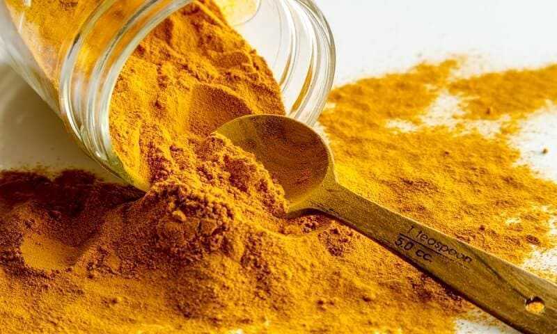 Close up of organic turmeric (curcuma) powder spilling out of glass jar with measuring spoon on white background