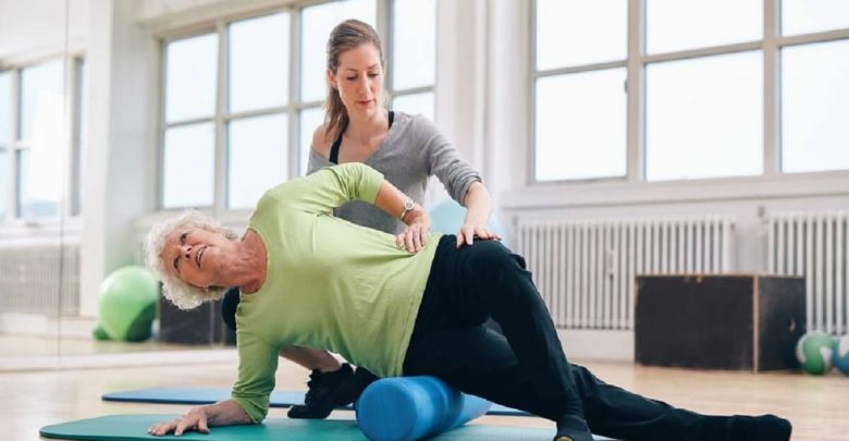 Female instructor helping senior woman using a foam roller for a myofascial release massage at gym